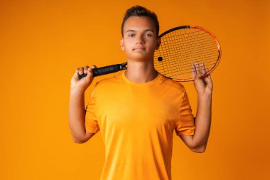 Turuncu arka planda raket tutan genç bir tenisçinin stüdyo çekimi.