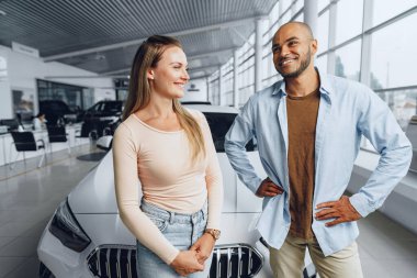 Kafkasyalı mutlu bir çift kadın ve Afro-Amerikalı bir adam arabalarının içinde yeni arabalarının yanında duruyorlar.