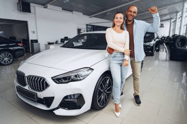 Kafkasyalı mutlu bir çift kadın ve Afro-Amerikalı bir adam arabalarının içinde yeni arabalarının yanında duruyorlar.