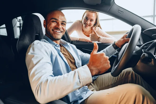 Joven pareja alegre mirando dentro de un coche nuevo que van a comprar en una tienda de coches — Foto de Stock