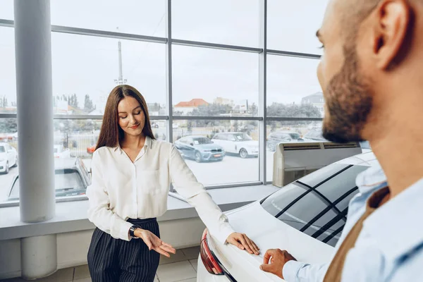 Ung attraktiv kvinna säljare i bil showroom visar en bil till sin manliga klient — Stockfoto