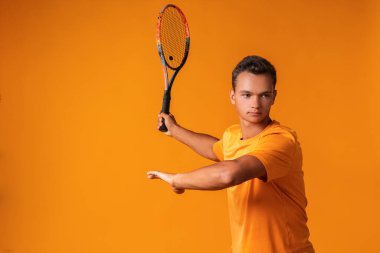 Turuncu arka planda raket tutan genç bir tenisçinin stüdyo çekimi.