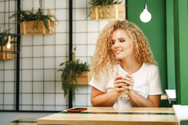 Kahve dükkanında masada oturan kıvırcık, genç bir kadın bir fincan kahveyle rahatlıyor.