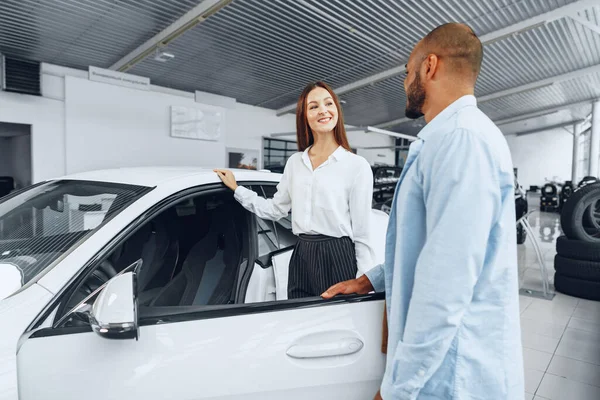 Junge attraktive Verkäuferin im Autohaus zeigt ihrem männlichen Kunden ein Auto — Stockfoto