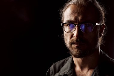 Gözlüklü, orta yaşlı bir adamın karanlık stüdyo portresi.