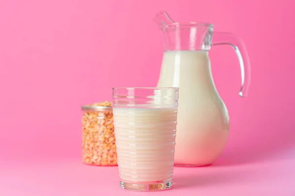 Oat milk in glassware and dry oat flakes on pink background