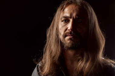 Uzun saçlı, erkeksi, orta yaşlı bir adam karanlıkta dikiliyor.