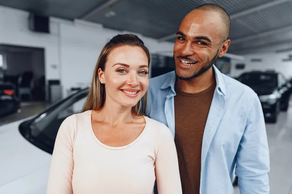 Szczęśliwa para kaukaska kobieta i afrykański amerykański mężczyzna stojący w pobliżu ich nowego samochodu wewnątrz salonu samochodowego — Zdjęcie stockowe