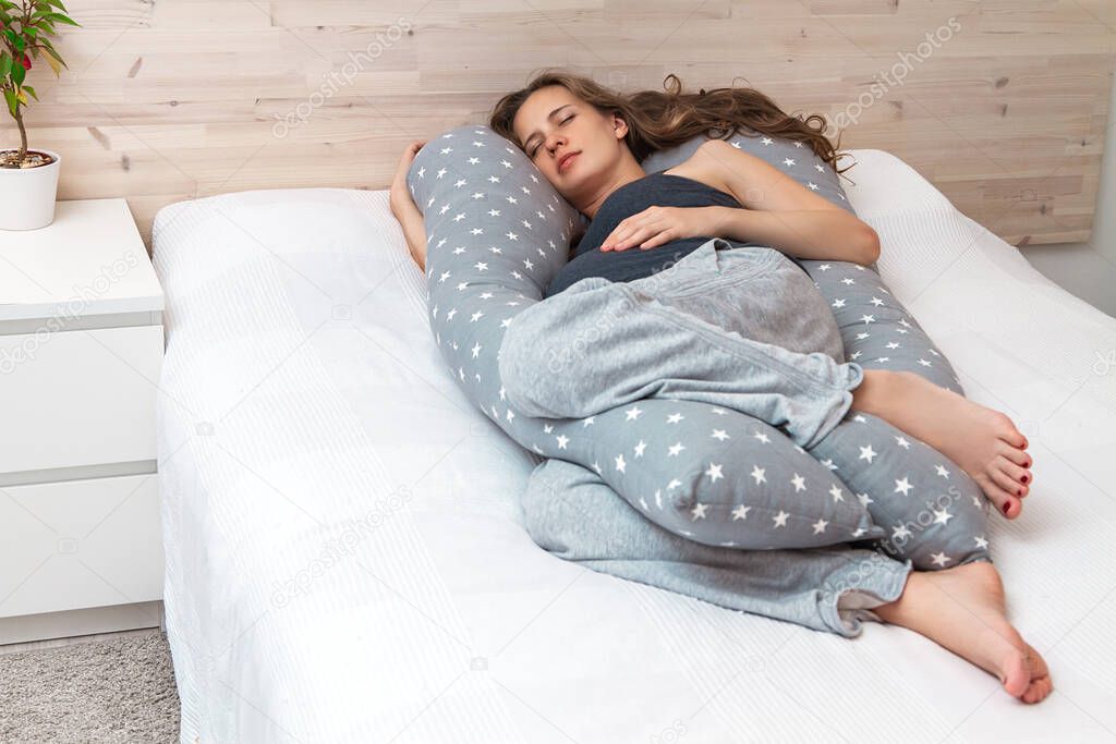 Beautiful sleeping pregnant woman with a big tummy with long curly hair and closed eyes, hugging a body pillow with her arm and legs and lying on the bed indoor.