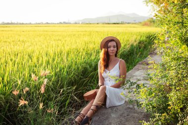 Kadın yol kenarında çantalı ve şapkalı, turist gibi oturuyor.