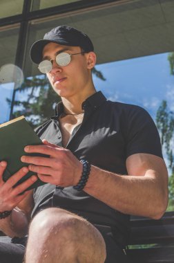 Avrupa adam, siyah bir gömlek ve bir cap City Park, bir kitap okumak'ta öğrenciyken yakınındaki nehrin