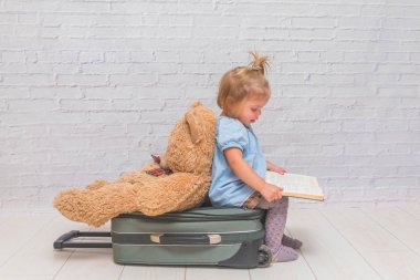 Kız, çocuk bir arka planda beyaz tuğla duvarın oturan bir sırtı bir kitapla oyuncak ayı için el valizle