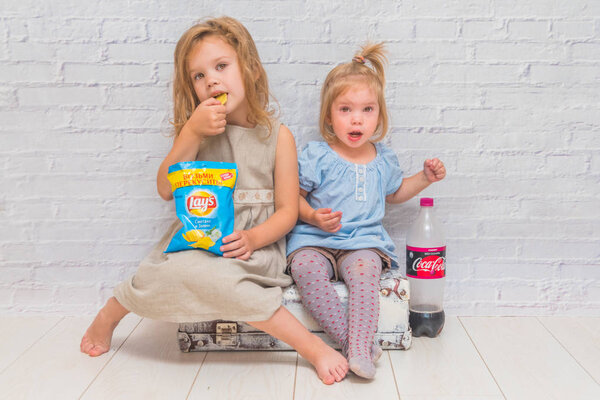 Russia, Nikolskoye, August 28, 2018-girls children sitting on a suitcase against a white brick wall eating chips, and drinking Coca-Cola