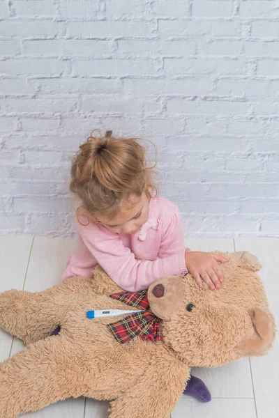 Девочка Ребенок Измеряет Температуру Игрушечного Медведя Прислонившись Стене Белых Брикетов — стоковое фото