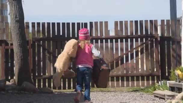 A menina, uma criança subiu sobre a cerca com um urso de brinquedo e uma mala — Vídeo de Stock