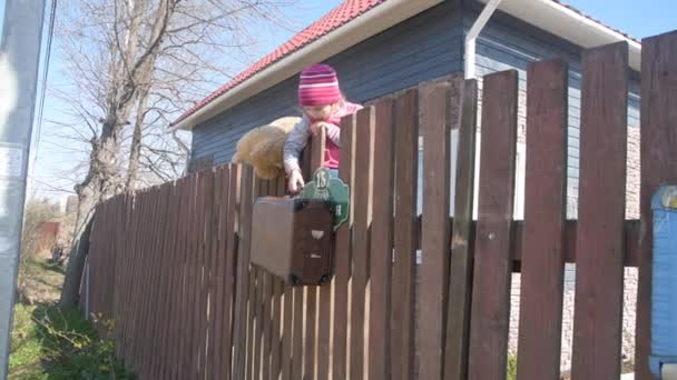 Το κορίτσι, ένα παιδί σκαρφάλωσε πάνω από το φράχτη με μια αρκούδα παιχνίδι και μια βαλίτσα — Αρχείο Βίντεο
