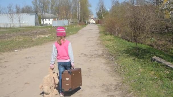 Девушка идет по дороге с чемоданом и игрушечным медведем в руке — стоковое видео