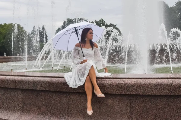 Piękna dziewczyna w białej sukni i parasol przy fontannie na — Zdjęcie stockowe