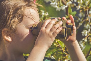 Çiçeklerin arka planında dürbünle bakan çocuk.