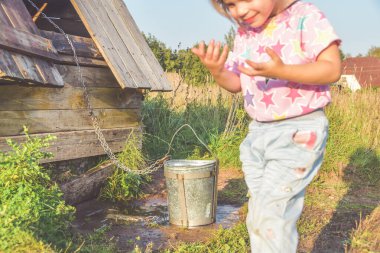 Güneşli yaz gününde kuyudaki bir çocuk kovayla su çeker ve metal bir bardaktan su içer.