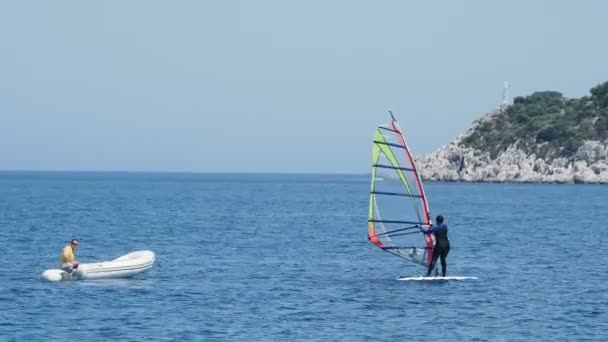 DEMRE, TURQUÍA - 16 de mayo de 2018. Mujer en traje de neopreno está levantando la vela de windsurf fuera del agua. El hombre en barco inflable con motor flota cerca de ella. Turquía . — Vídeos de Stock