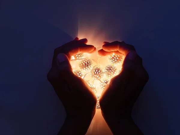 Hands with light bulbs in shape of heart. Metal light bulbs with delicate pattern shine in dark. Holiday decoration with magic light reflections.