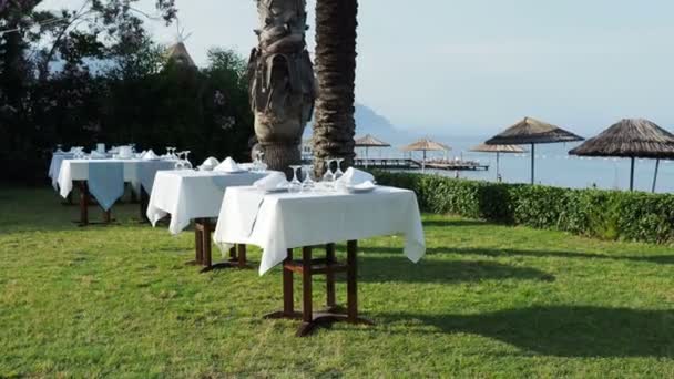 Mesas, servidas para la cena en césped verde. Comida mediterránea al aire libre bajo palmeras. Cena a orillas del mar . — Vídeos de Stock