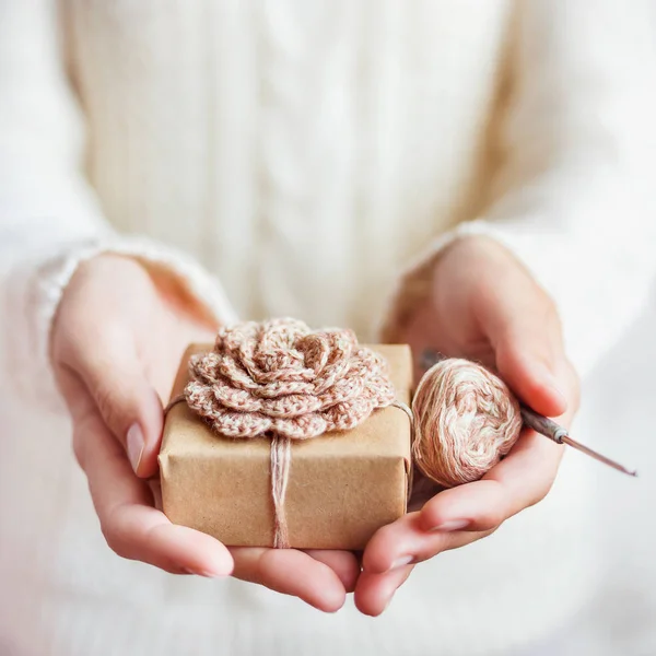 Woman in white knitted sweater holding a present wrapped in craft paper with hand made crocheted flower. DIY Cristmas or New Year gift.