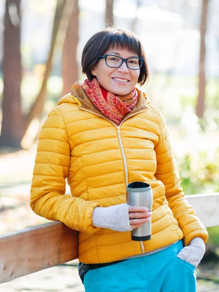 Happy wide smiling women in bright yellow jacket is holding thermos mug. Hot tea or other beverage on cool autumn day.