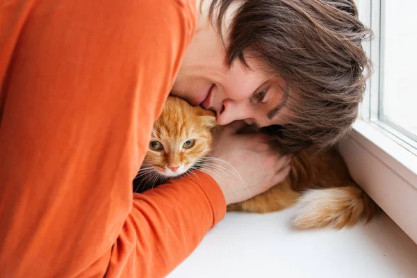 Man in orange sweatshirt is hugging cute ginger cat. Pet adoption. Ex-stray cat at home. Fluffy pet and it\'s master.