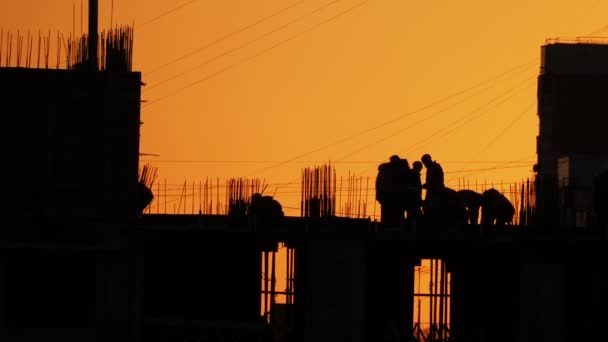 Byggandet av bostadshus. Byggare går på oavslutade golvet med utstickande beslag. Siluetter av arbetstagare på bakgrunden av en orange solnedgång. — Stockvideo