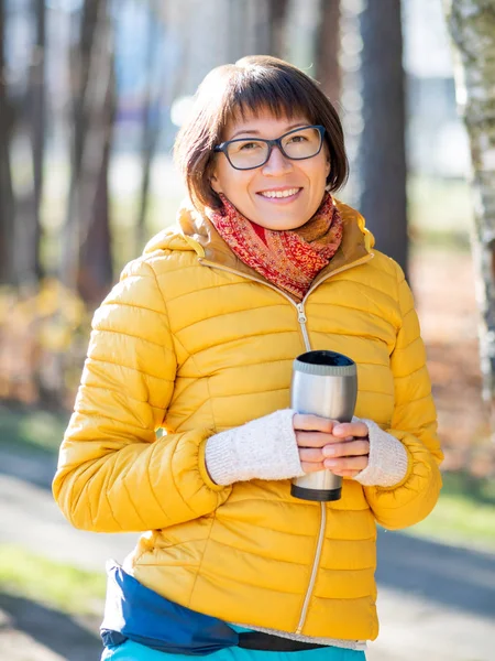 Happy wide smiling women in bright yellow jacket is holding thermos mug. Hot tea or other beverage on cool autumn day.