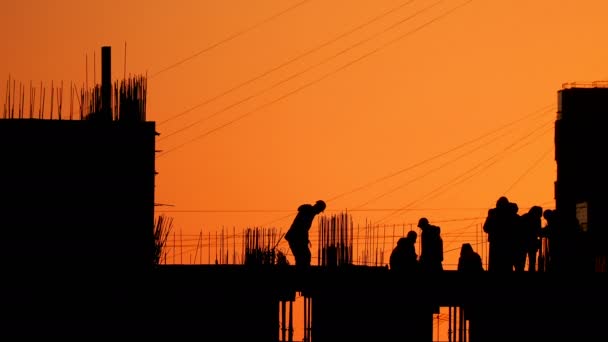 Construcción de un edificio residencial. Los constructores van en el piso sin terminar con accesorios sobresalientes. Siluetas de los trabajadores en el fondo de una puesta de sol naranja . — Vídeo de stock