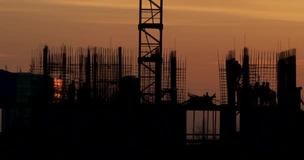 Bau von Wohngebäuden. Bauarbeiter gehen mit überstehenden Beschlägen auf den Rohboden. Silhouetten von Arbeitern vor einem orangefarbenen Sonnenuntergang. — Stockvideo