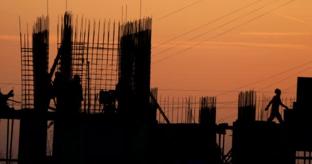 Bau von Wohngebäuden. Bauarbeiter gehen mit überstehenden Beschlägen auf den Rohboden. Silhouetten von Arbeitern vor einem orangefarbenen Sonnenuntergang. — Stockvideo