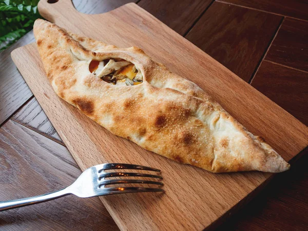 Baked bun in the shape of a boat with mozzarella cheese, cheddar cheese and oyster mushrooms. Pastry on wooden chopping board.