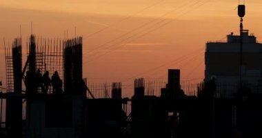 Konut bina inşaatı. İnşaatçılar bağlantı parçaları çıkıntılı bitmemiş yere gidin. Arka planda bir turuncu günbatımı işçilerin siluetleri.