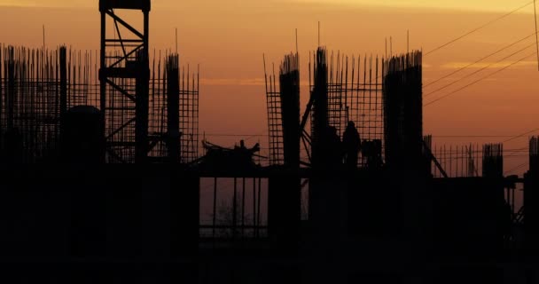 Construcción de un edificio residencial. Los constructores van en el piso sin terminar con accesorios sobresalientes. Siluetas de los trabajadores en el fondo de una puesta de sol naranja . — Vídeo de stock