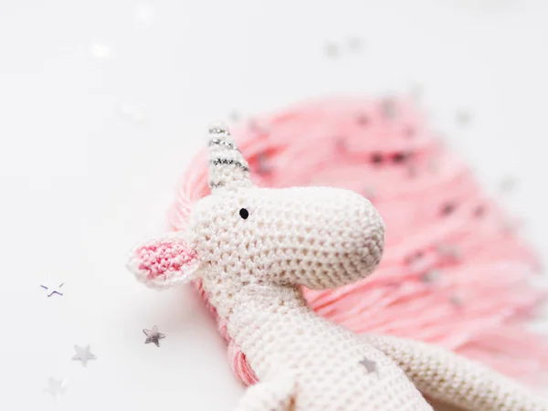 Lindo unicornio de hadas con una melena rosa y una cola hecha de hilos. Juguete hecho a mano de ganchillo sobre fondo blanco con confeti estrellas de plata. Criatura de moda, símbolo de magia y milagros . — Foto de Stock