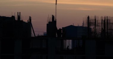 Konut bina inşaatı. İnşaatçılar bağlantı parçaları çıkıntılı bitmemiş yere gidin. Arka planda bir turuncu günbatımı işçilerin siluetleri.