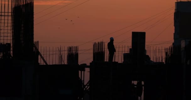 Construction d'un bâtiment résidentiel. Les constructeurs vont sur le plancher inachevé avec des raccords saillants. Silhouettes de travailleurs sur le fond d'un coucher de soleil orange . — Video