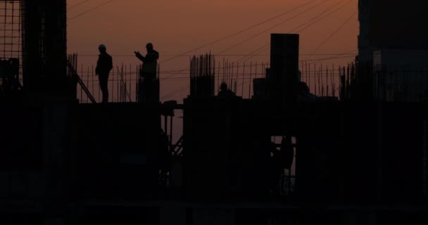 Строительство жилого дома. Строители идут по недостроенному этажу с проторенной арматурой. Силуэты рабочих на фоне оранжевого заката . — стоковое видео