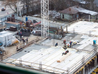 Eski ahşap evler arasında şantiyede havadan görünüm. Şantiyedeki işçiler tatilde. Beton takviye ve inşaat ekipmanları arasında turuncu üniformalı inşaatçılar. Rusya.