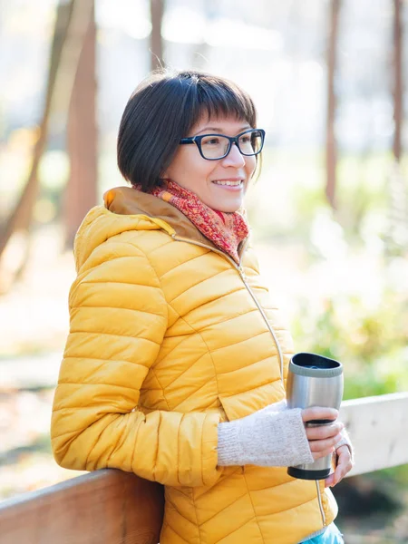 Gelukkig breed glimlachende vrouwen in heldere gele jas houdt thermos beker. Hete thee of andere drank op cool herfstdag. — Stockfoto