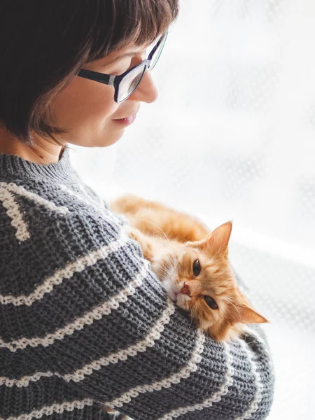 Mignon chat roux somnolant sur les bras de la femme. Femme souriante en pull tricoté gris caressant son animal de compagnie moelleux. Maison confortable . — Photo