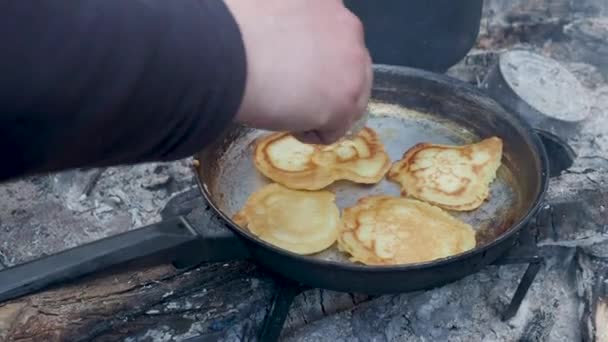 O turista está cozinhando panquecas na frigideira sobre o fogo. Fogueira na floresta . — Vídeo de Stock