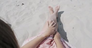Kumlu sahilde dinlenen kadın. Rüzgar pembe etek açık kumaş sallar. Kadın tabaklanmış elleriyle giysileri tutar. Omzunun arkasından çekim.