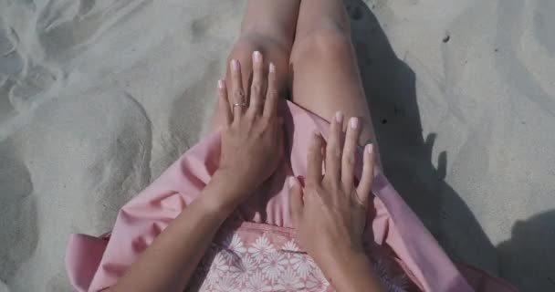 Mulher descansando na praia de areia. Vento balança tecido leve de saia rosa. Mulher segura a roupa com as mãos bronzeadas. Tiro por trás do ombro . — Vídeo de Stock