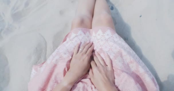Mulher descansando na praia de areia. Vento balança tecido leve de saia rosa. Mulher segura a roupa com as mãos bronzeadas. Tiro por trás do ombro . — Vídeo de Stock