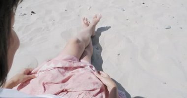 Kumlu sahilde dinlenen kadın. Rüzgar pembe etek açık kumaş sallar. Kadın tabaklanmış elleriyle giysileri tutar. Omzunun arkasından çekim.